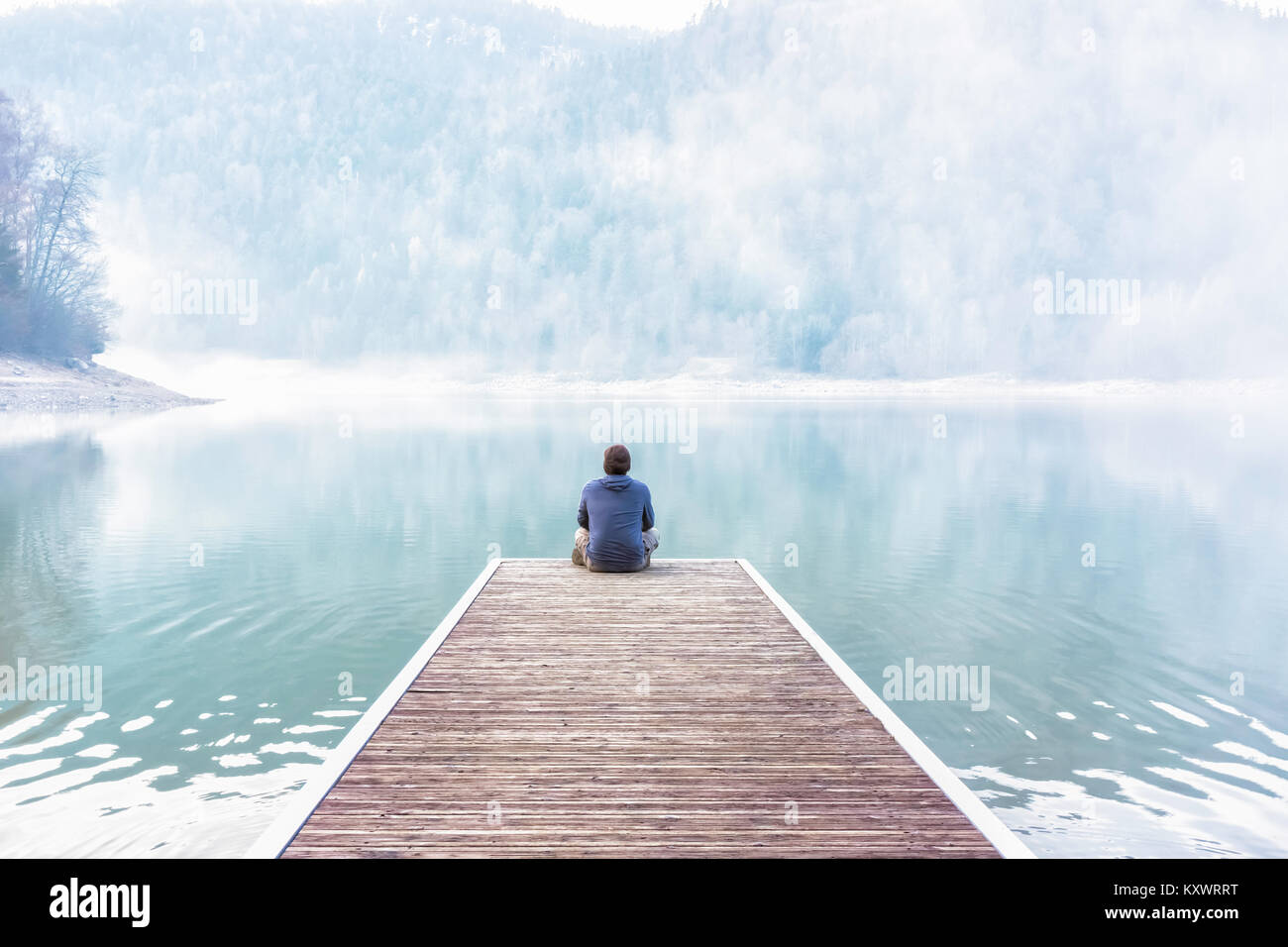 Jeune homme assis et méditant sur le quai d'un lac en un jour brumeux de l'hiver, Vosges, France Banque D'Images