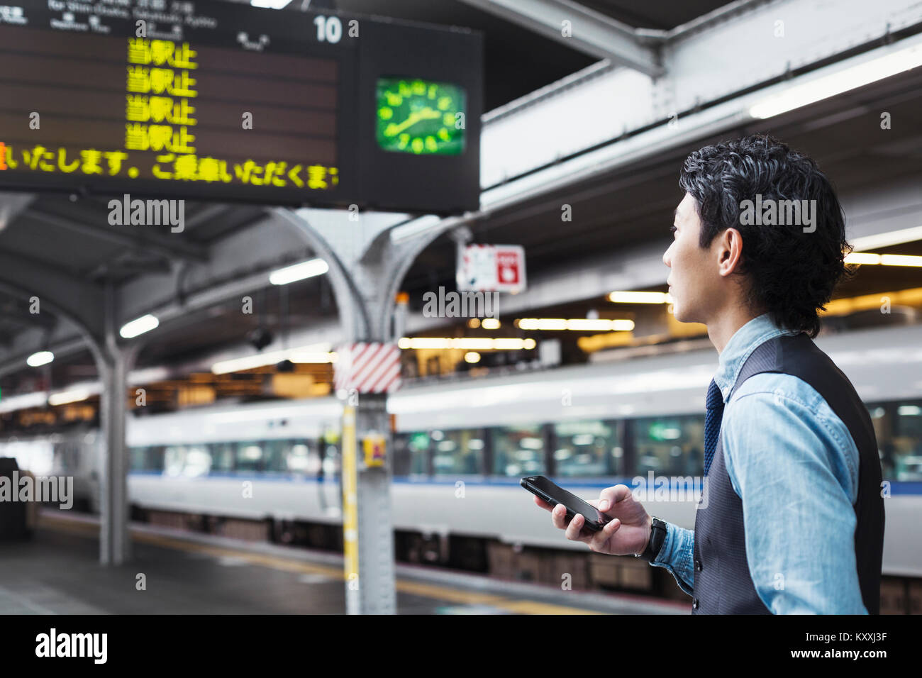 Businessman wearing blue shirt et gilet debout sur la plate-forme de la gare, holding mobile phone. Banque D'Images