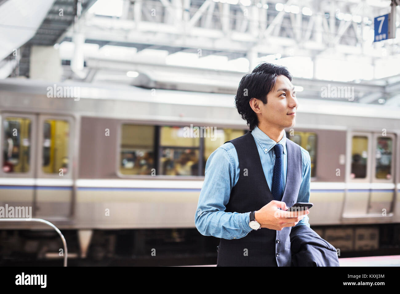 Businessman wearing blue shirt et gilet debout sur la plate-forme de la gare, holding mobile phone. Banque D'Images