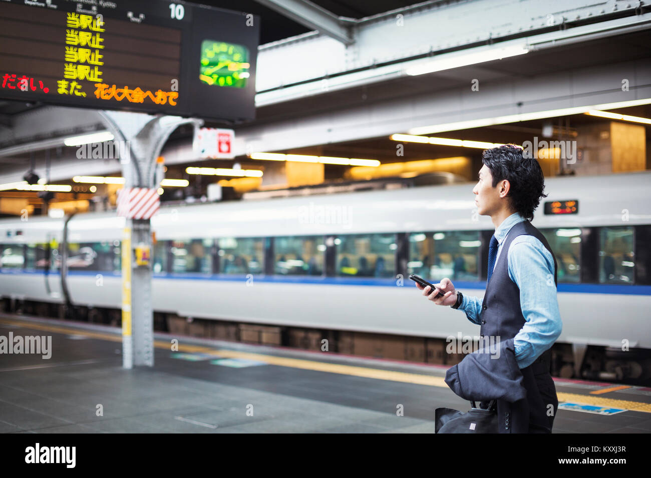 Businessman wearing blue shirt et gilet debout sur la plate-forme de la gare, holding mobile phone. Banque D'Images