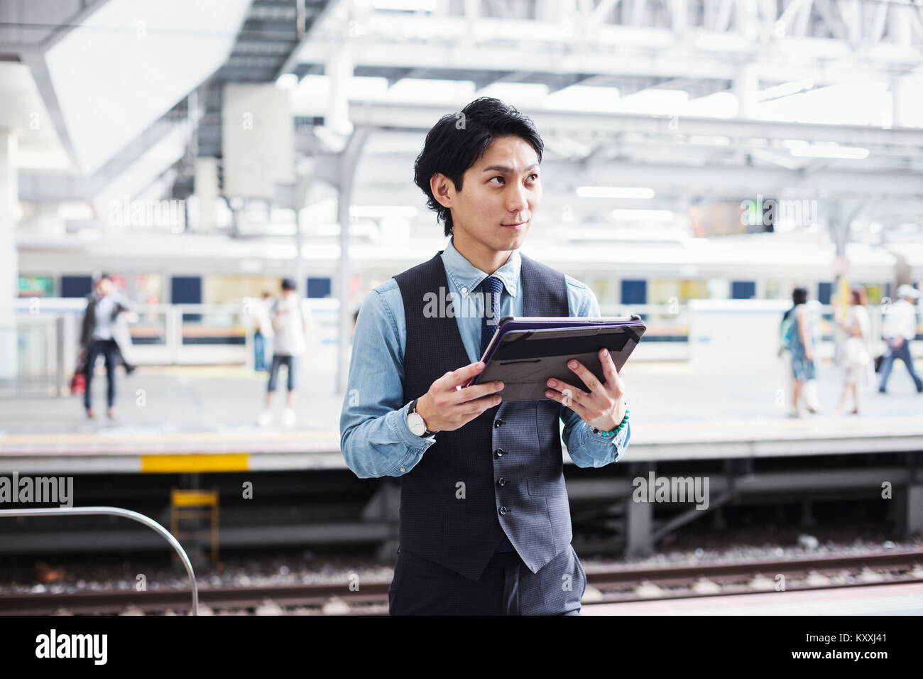Businessman wearing blue shirt et gilet debout sur la plate-forme de la gare, holding digital tablet. Banque D'Images