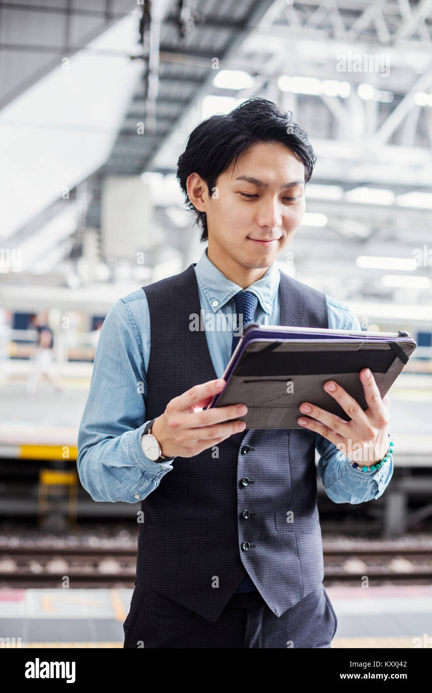Businessman wearing blue shirt et gilet debout sur la plate-forme de la gare, holding digital tablet. Banque D'Images