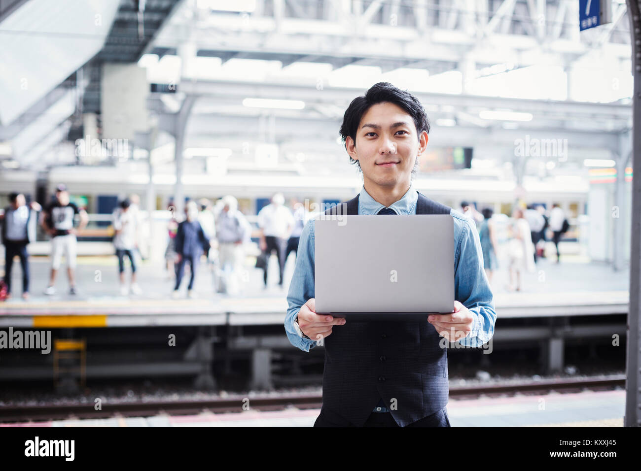 Businessman wearing blue shirt et gilet debout sur la plate-forme de la gare, holding laptop, looking at camera. Banque D'Images