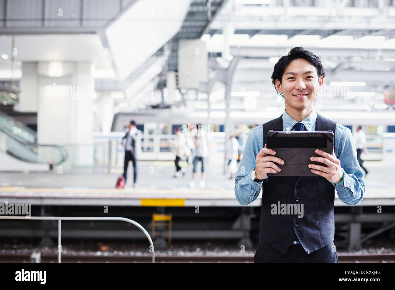 Businessman wearing blue shirt et gilet debout sur la plate-forme de la gare, holding digital tablet, looking at camera. Banque D'Images