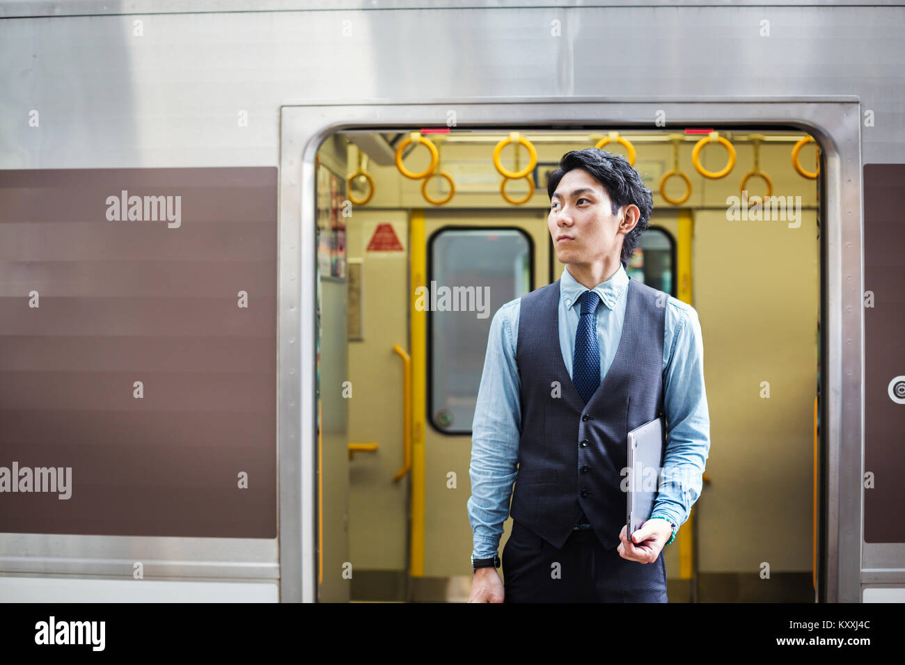 Businessman wearing blue shirt et gilet debout sur la plate-forme de la gare, holding laptop. Banque D'Images