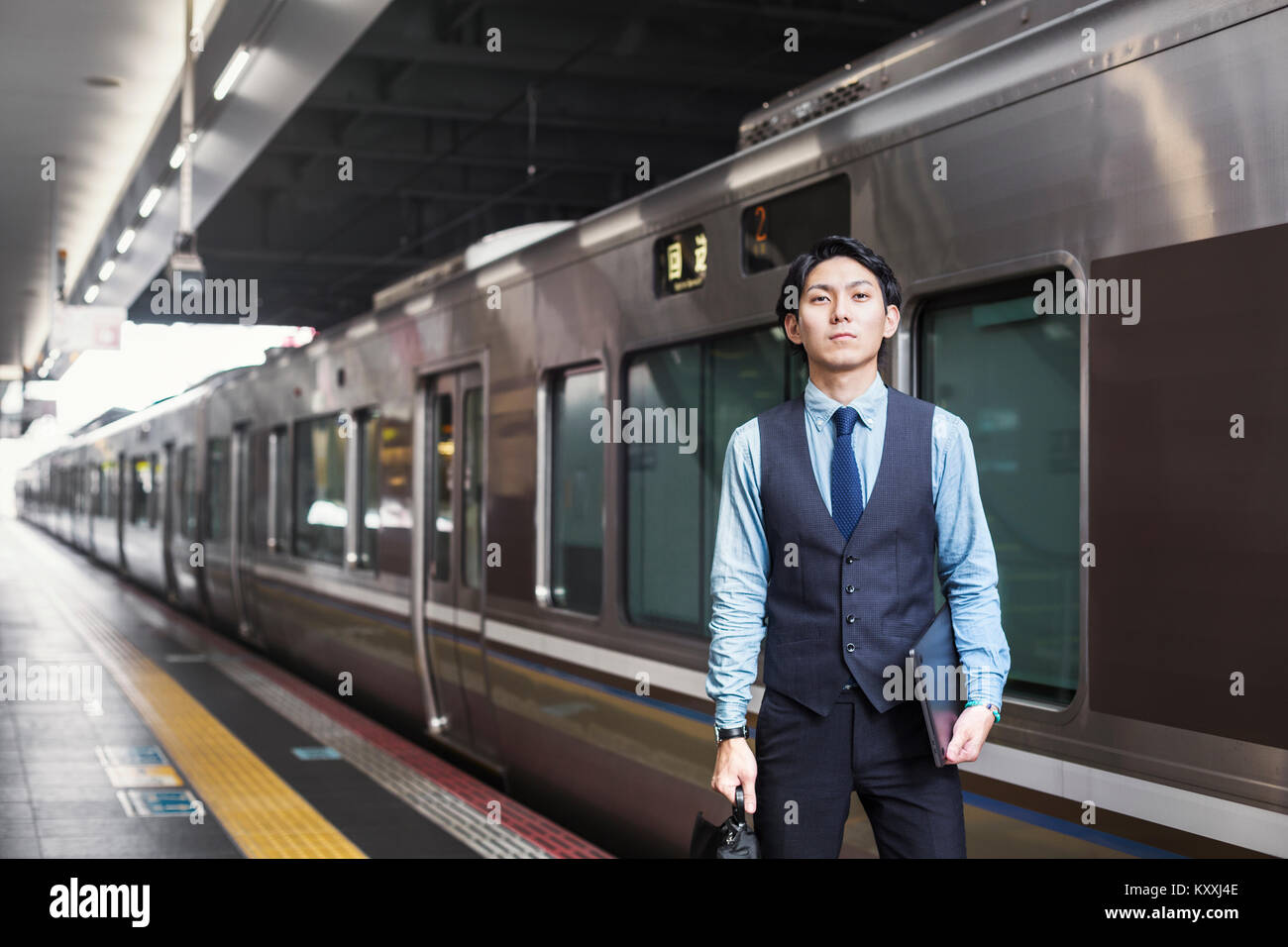 Businessman wearing blue shirt et gilet debout sur la plate-forme de la gare, holding digital tablet et porte-documents. Banque D'Images