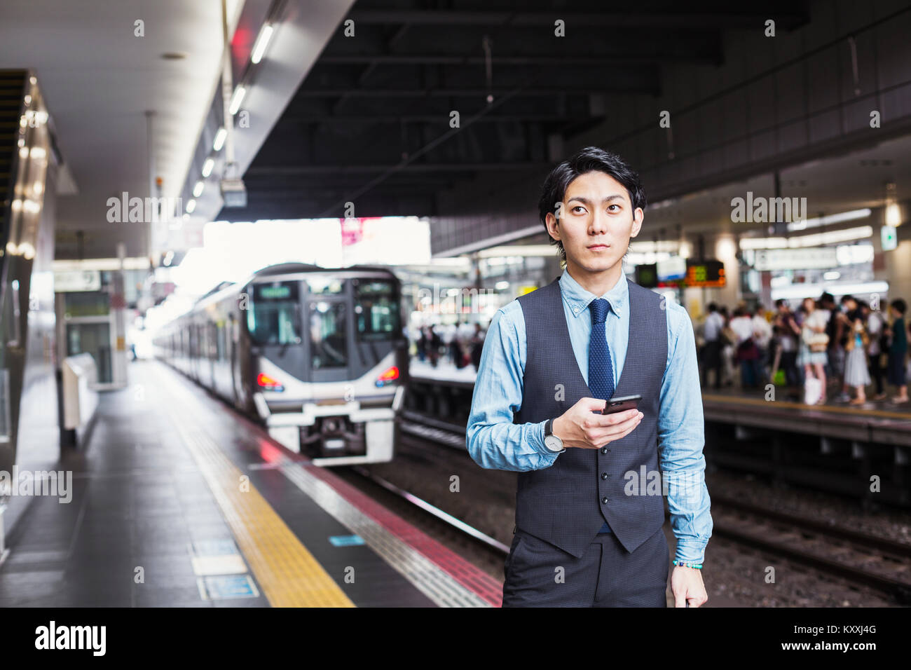 Businessman wearing blue shirt et gilet debout sur la plate-forme de la gare, holding mobile phone. Banque D'Images