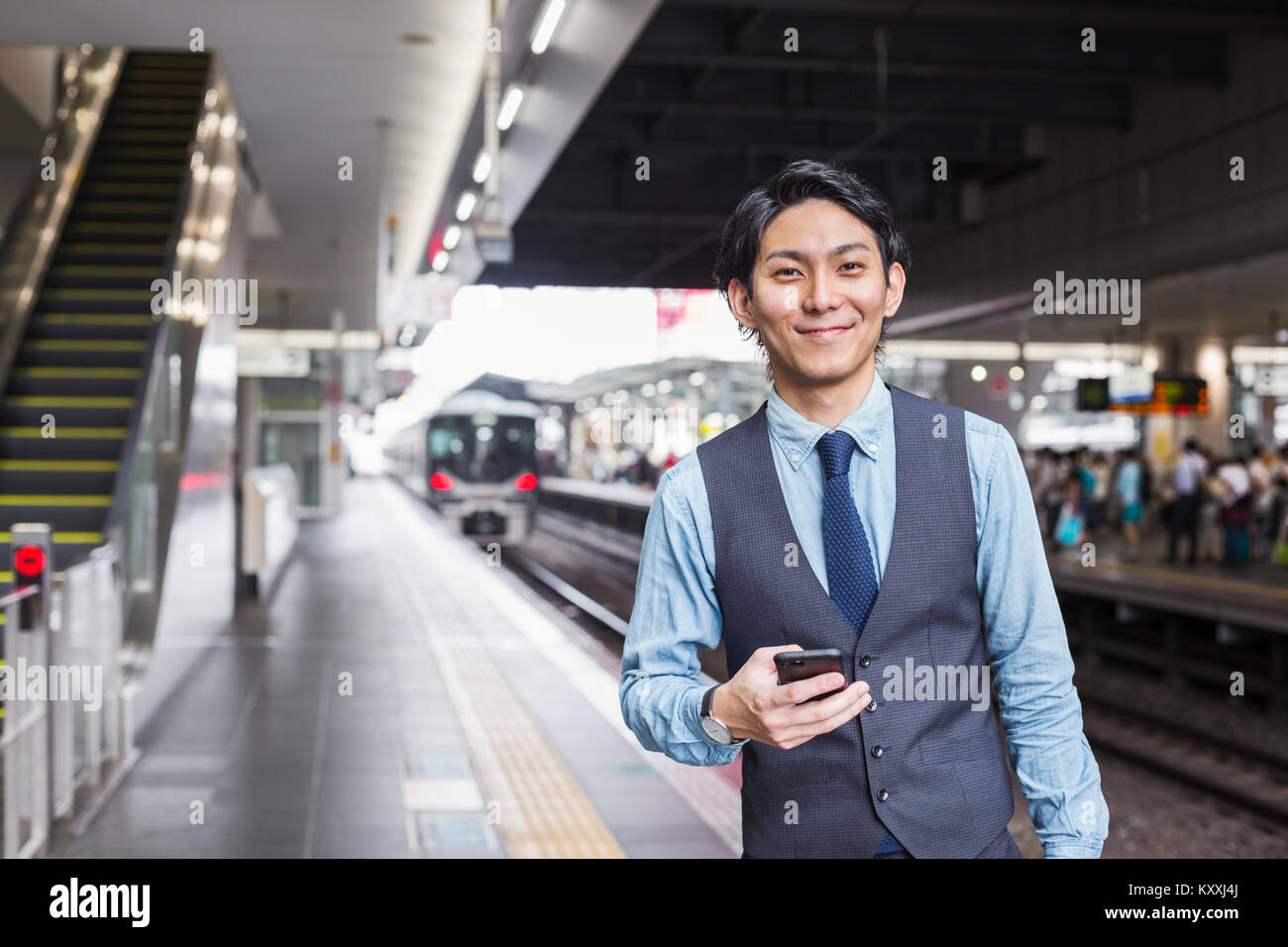 Businessman wearing blue shirt et gilet debout sur la plate-forme de la gare, holding mobile phone. Banque D'Images