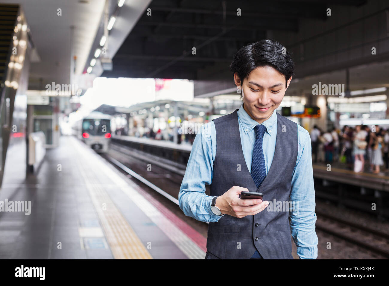 Businessman wearing blue shirt et gilet debout sur la plate-forme de la gare, holding mobile phone. Banque D'Images