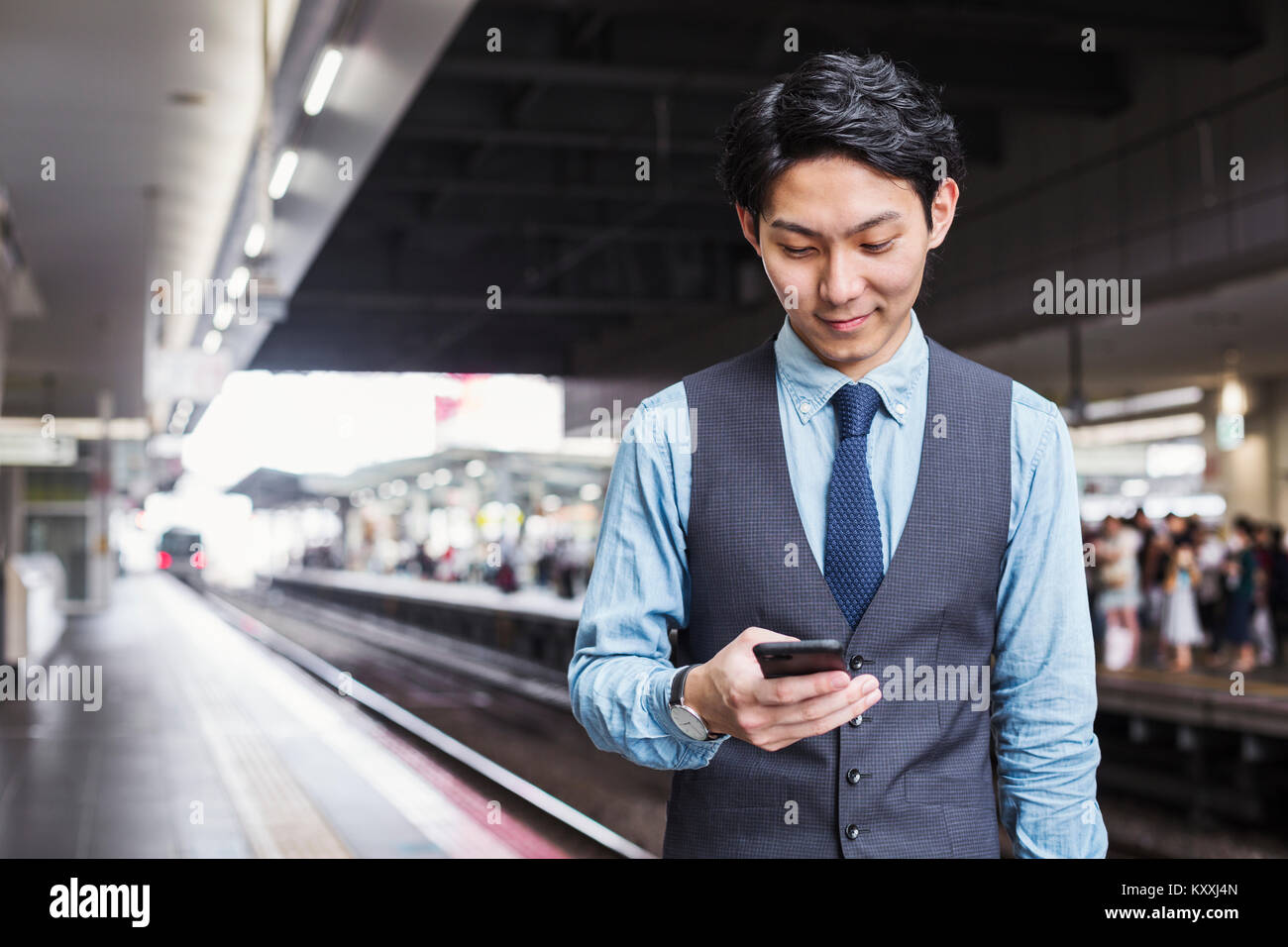 Businessman wearing blue shirt et gilet debout sur la plate-forme de la gare, holding mobile phone. Banque D'Images