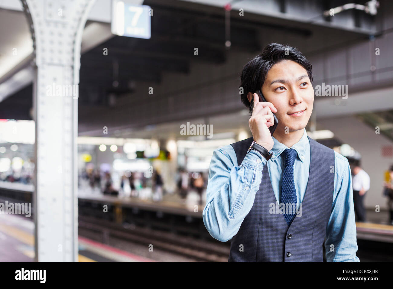 Businessman wearing blue shirt et gilet debout sur la plate-forme de la gare, talking on mobile phone. Banque D'Images