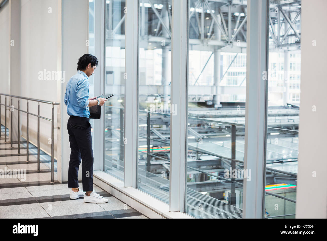 Businessman wearing blue shirt debout à l'intérieur par un mur de verre, holding digital tablet. Banque D'Images