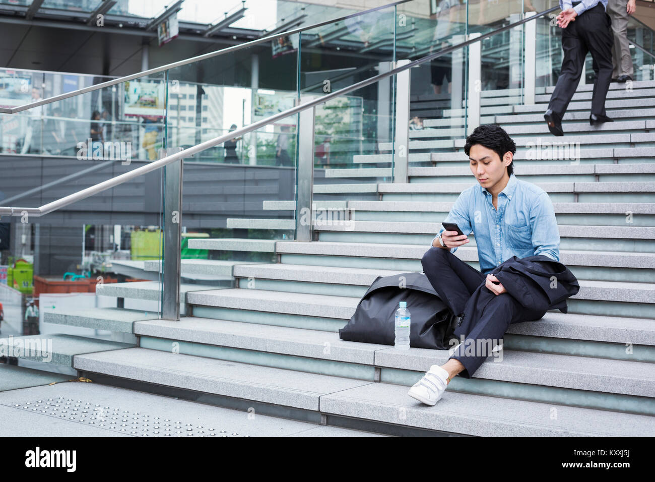 Businessman wearing blue shirt assis dehors sur les marches, looking at mobile phone. Banque D'Images