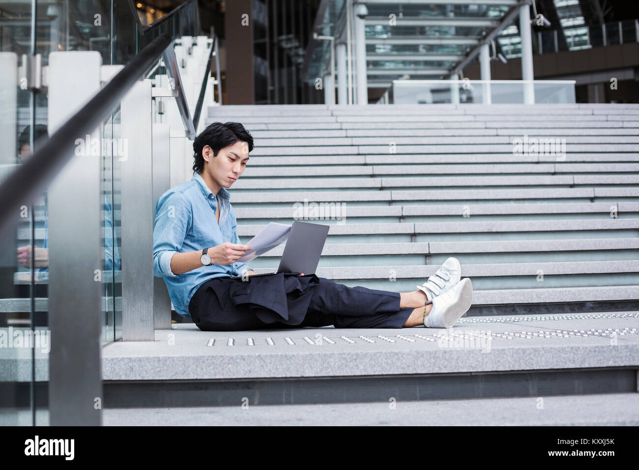 Businessman wearing blue shirt assis dehors sur les marches, holding documents, ordinateur portable sur ses genoux. Banque D'Images