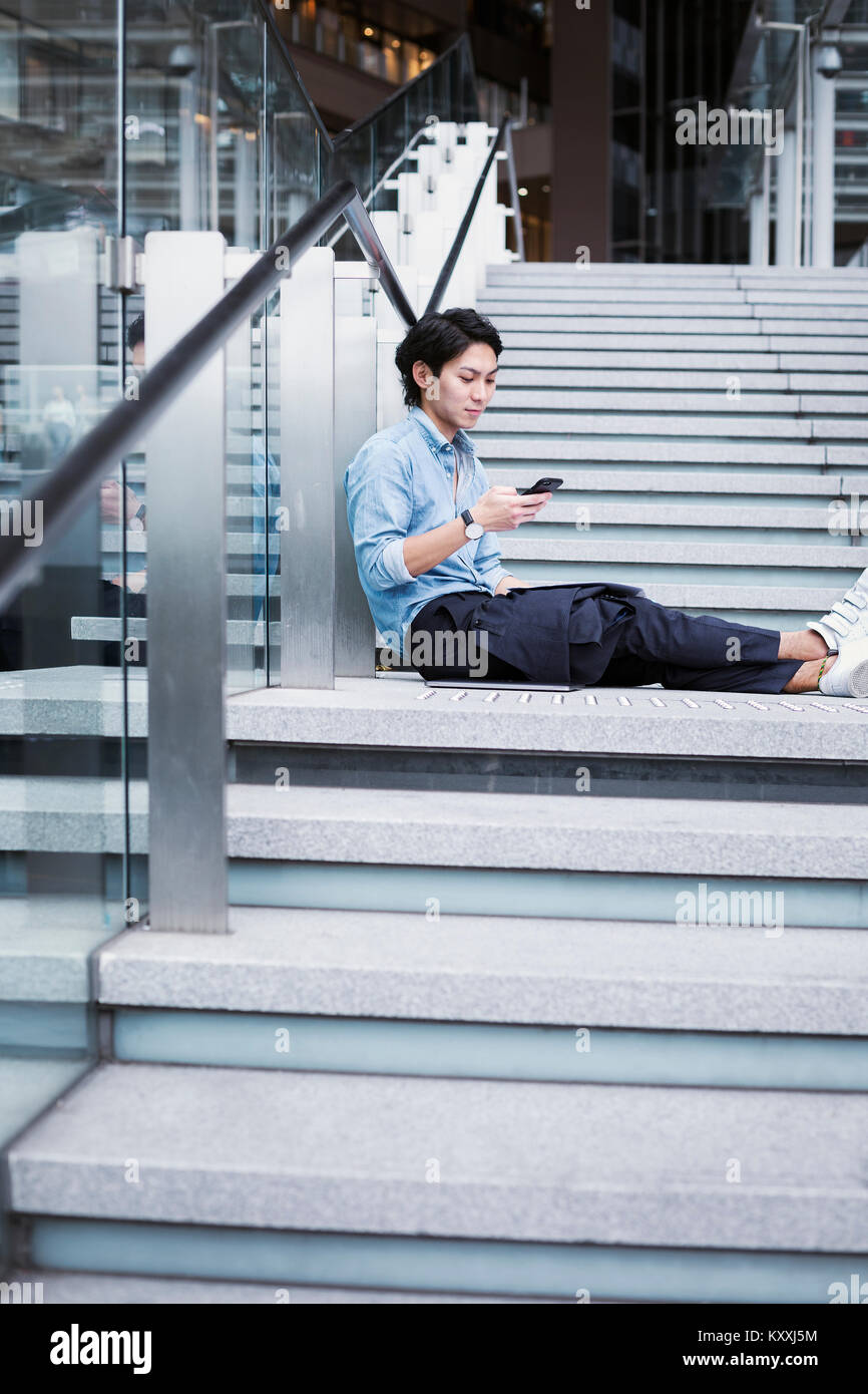 Businessman wearing blue shirt assis dehors sur les marches, tenant le téléphone mobile. Banque D'Images