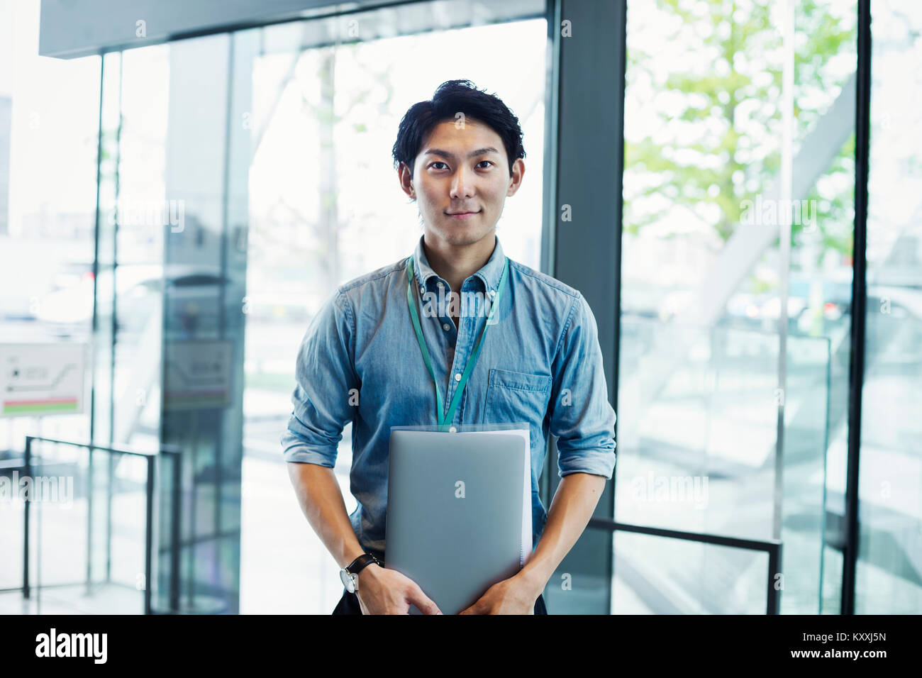 Businessman wearing blue shirt debout à l'intérieur par un mur de verre, holding laptop, looking at camera. Banque D'Images