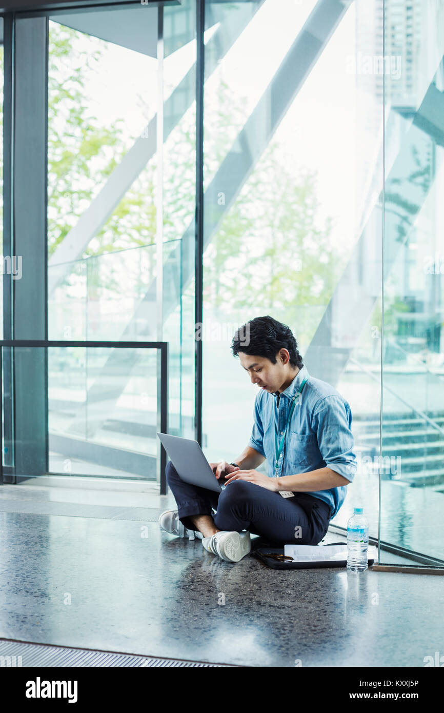 Businessman wearing blue shirt assis sur le plancher à l'intérieur, appuyé contre la paroi de verre, travailler sur un ordinateur portable. Banque D'Images