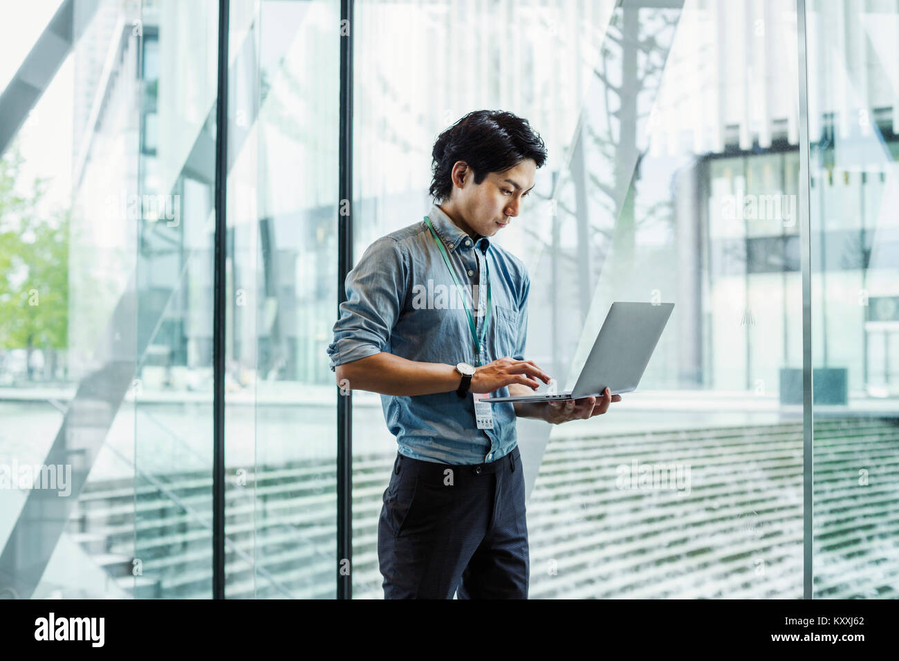 Businessman wearing blue shirt debout à l'intérieur par un mur de verre, holding laptop. Banque D'Images