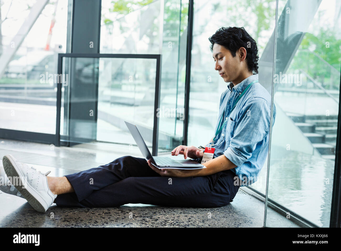 Businessman wearing blue shirt assis sur le plancher à l'intérieur, appuyé contre la paroi de verre, travailler sur un ordinateur portable. Banque D'Images