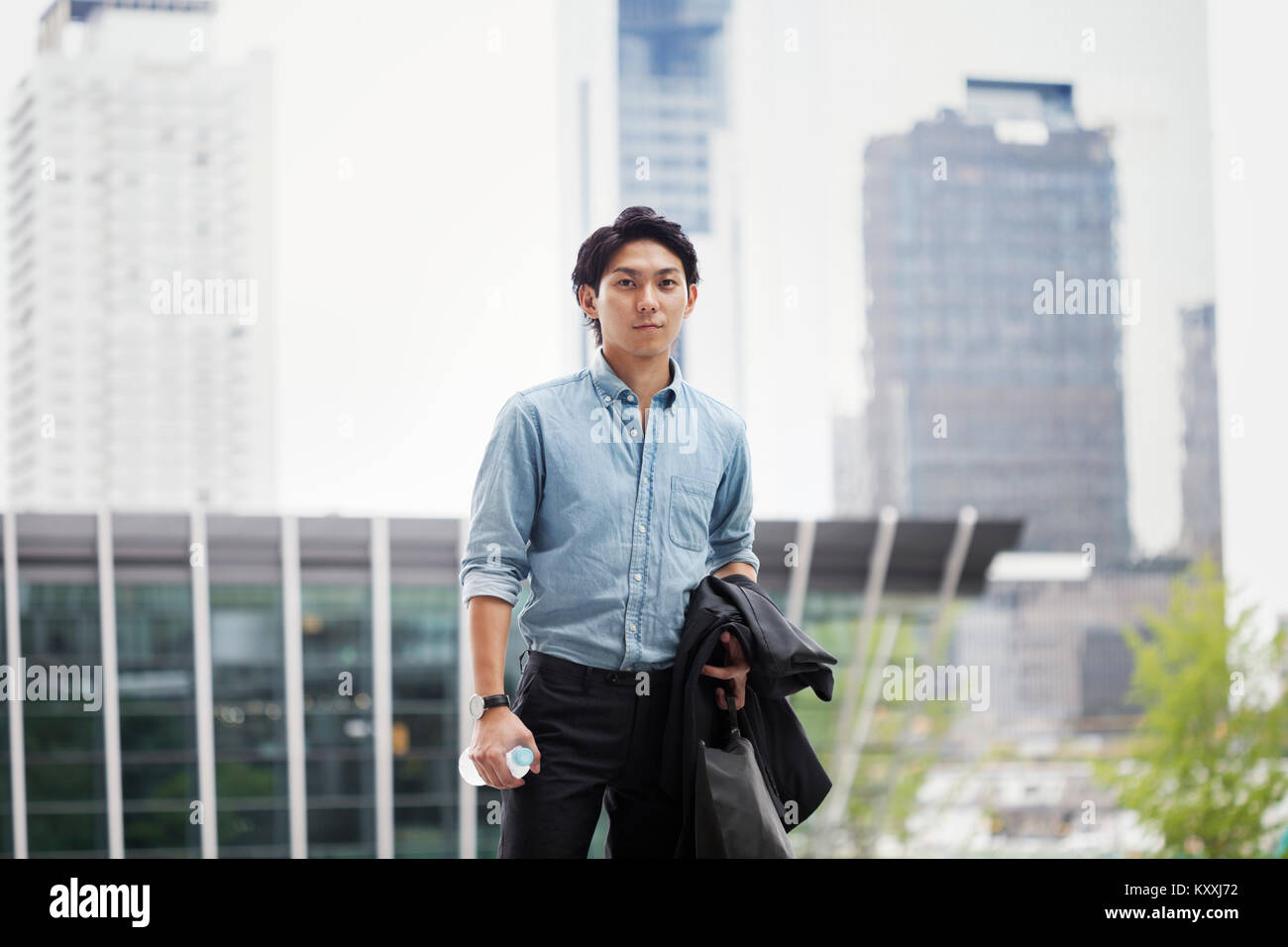 Businessman wearing blue shirt standing outdoors, holding jacket, serviette et bouteille d'eau. Banque D'Images