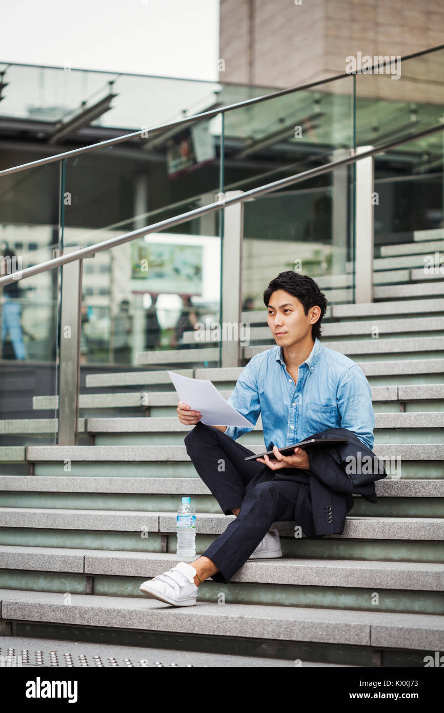 Businessman wearing blue shirt assis dehors sur les marches, holding digital tablet et papiers. Banque D'Images