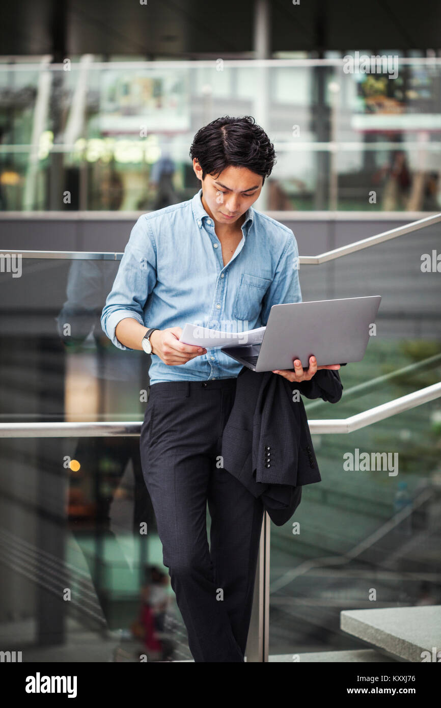 Businessman wearing blue shirt standing outdoors, holding laptop et papiers, appuyé contre rampe en verre. Banque D'Images