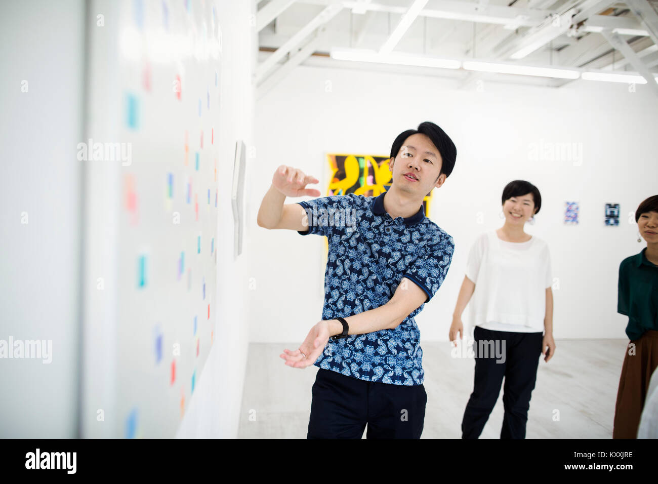 Man wearing blue shirt debout à côté de la peinture moderne sur le mur blanc dans la galerie d'art, deux femmes se tenant debout en arrière-plan.. Banque D'Images