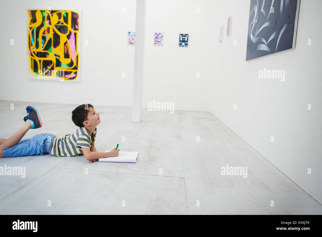 Garçon aux cheveux noirs courts lying on floor en galerie d'art avec un stylo et du papier, à la recherche de la peinture moderne. Banque D'Images