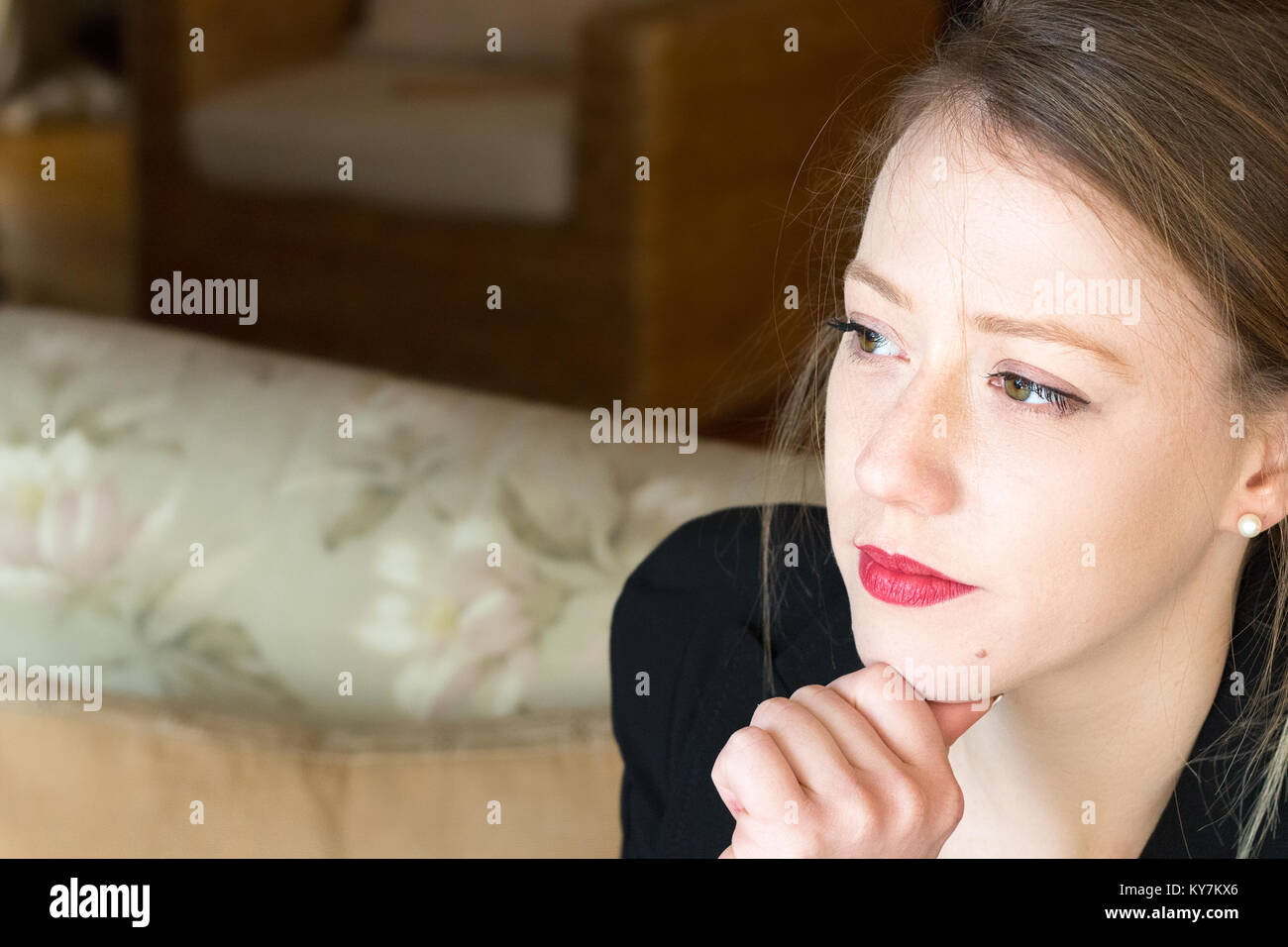 Portrait d'une jeune femme assise à la maison avec son pouce sur son menton, pensive, en pensant à la suite. Banque D'Images