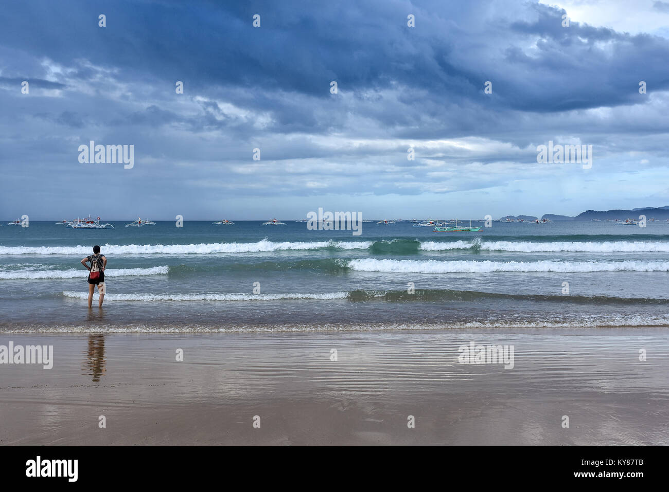 Une jeune femme debout à la cheville dans une mer tropicale à la pompe traditionnelle des Philippines à bateaux et le passage des nuées d'orage. Banque D'Images