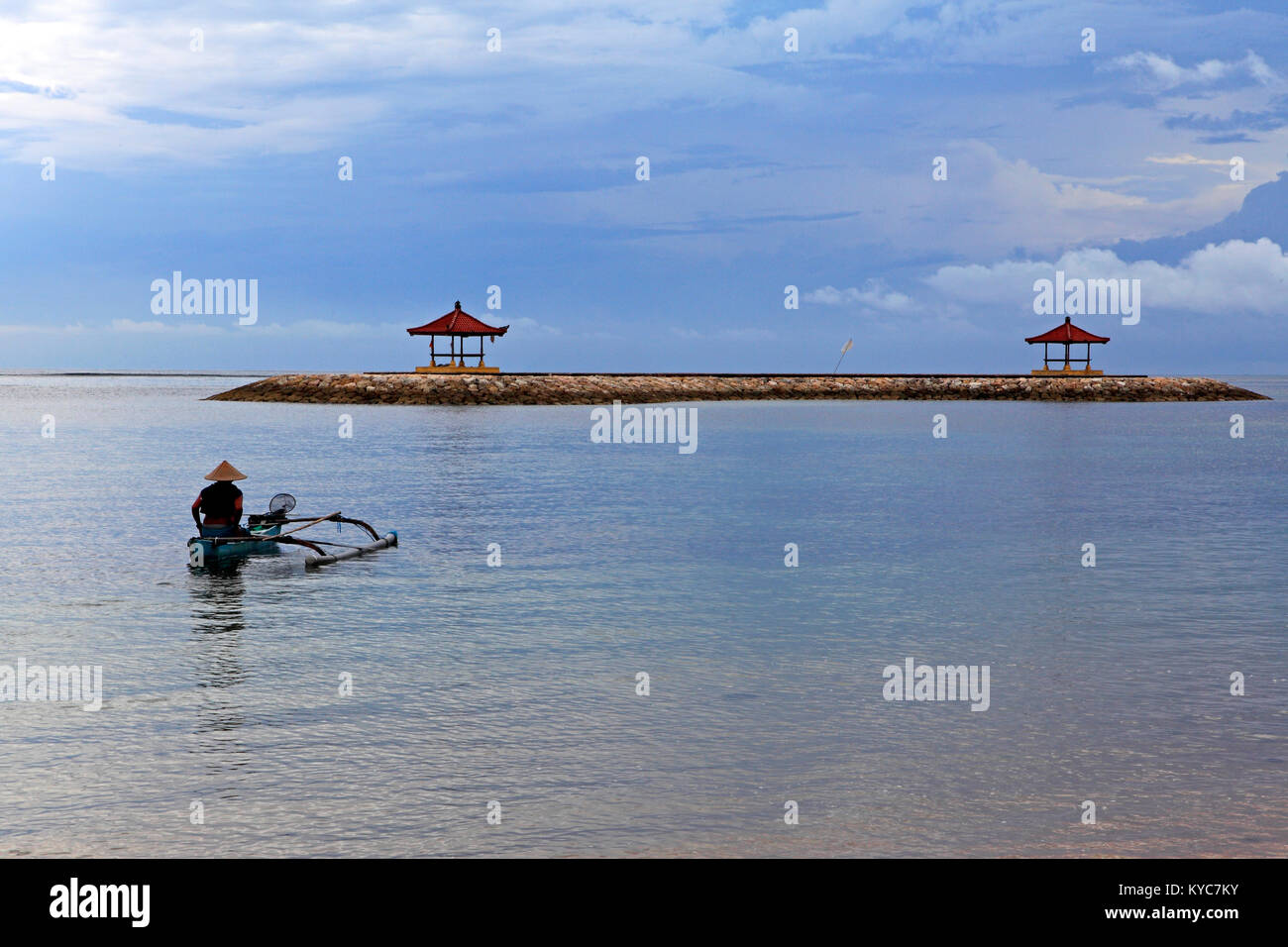Pêcheur balinais dans un outrigger bateau en bois, portant un chapeau conique, palettes dans la lagune à la plage de Sanur en face de deux pagodes. Bali. Banque D'Images