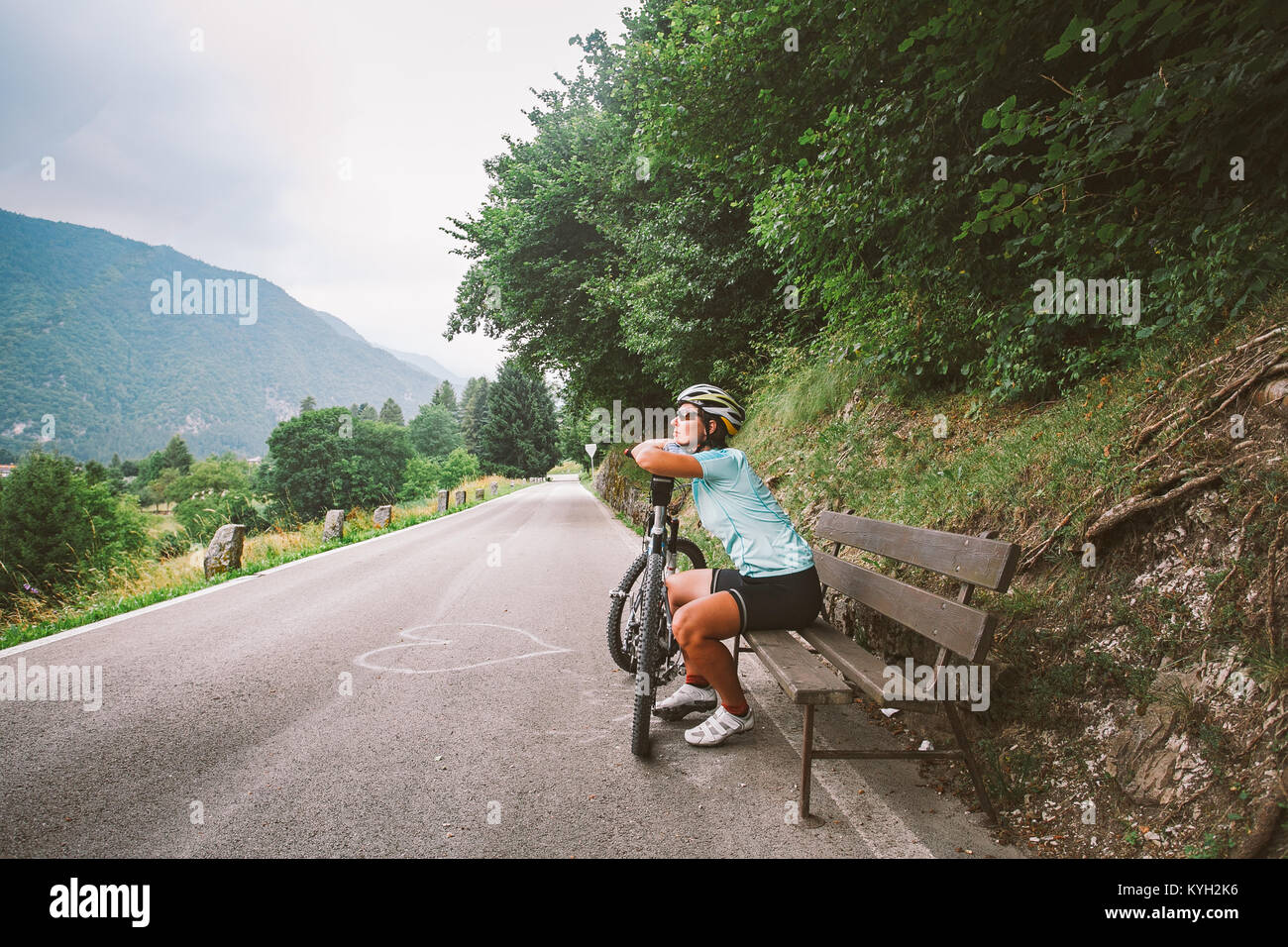 Jeune fille dans un casque et des vêtements de sport et de rêves de repos assis regardant dans la distance sur un banc en bois sur une piste cyclable en Italie en moun Banque D'Images