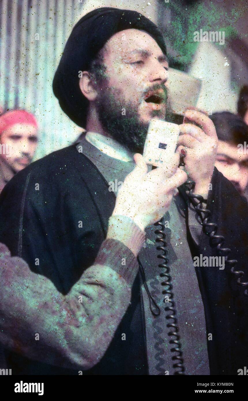 Vue d'un homme habillé en robe de bureau traditionnels et turban noir, parlant dans un micro à un rassemblement à l'Iran, mars, 1983. Le bras d'une personne tend la main tenant un enregistreur de voix du côté gauche du châssis. D'importants dégâts sur l'original. () Banque D'Images