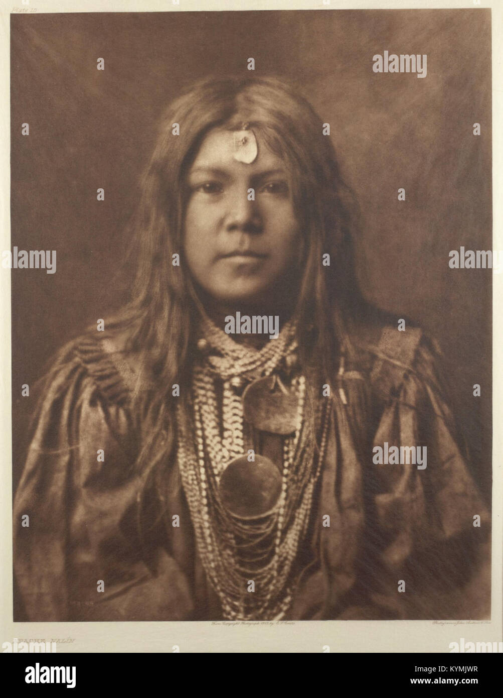 Photographie par Edward S. Curtis d'une fille amérindienne Apache ornée de perles traditionnelles, collier de coquillages et coiffe à franges. Les travaux de Curtis au début du XXe siècle documentent les cultures amérindiennes. Banque D'Images