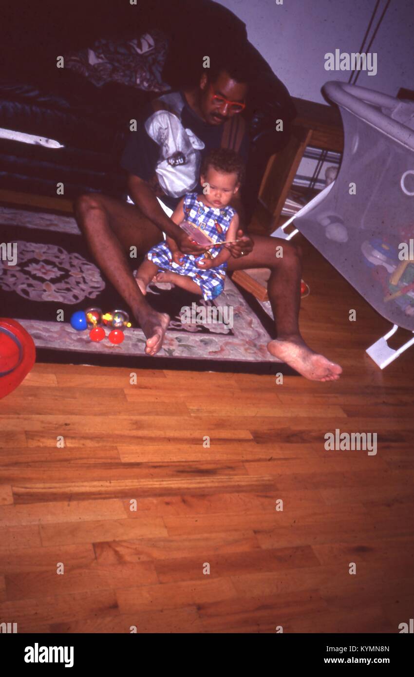 Vue d'un African American man lire un livre à une jeune africaine-américaine, de l'enfant, juillet 1995. La petite fille et l'homme s'asseoir ensemble sur le plancher d'une salle de séjour de la famille, avec un canapé et d'un parc à l'arrière-plan. () Banque D'Images