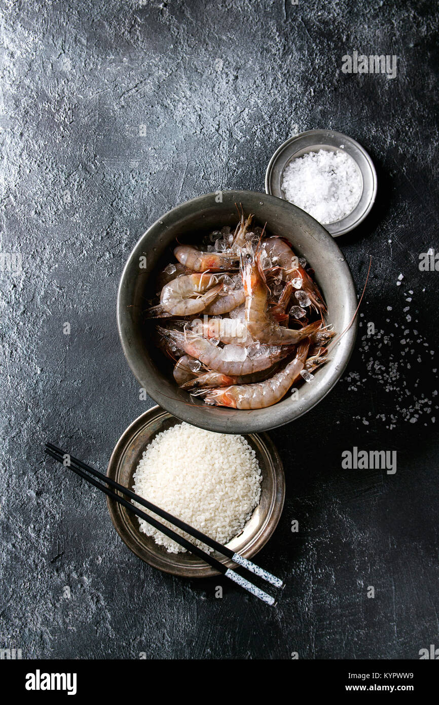 Crevettes non cuites fraîches entières, les crevettes sur la glace dans un bol en métal vintage avec des ingrédients ci-dessus. Le sel de mer, le riz, l'ail, baguettes sur tex noir Banque D'Images