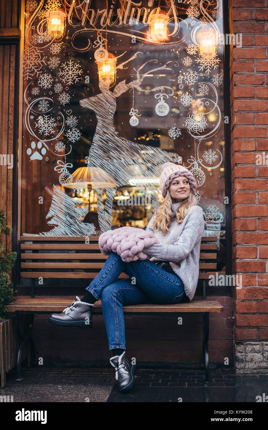 Young blonde woman in hat tricoté rose fumé, col en tricot et les blue-jeans faire poser sur un banc en bois avec noël cafe-fenêtre sur backgrou Banque D'Images