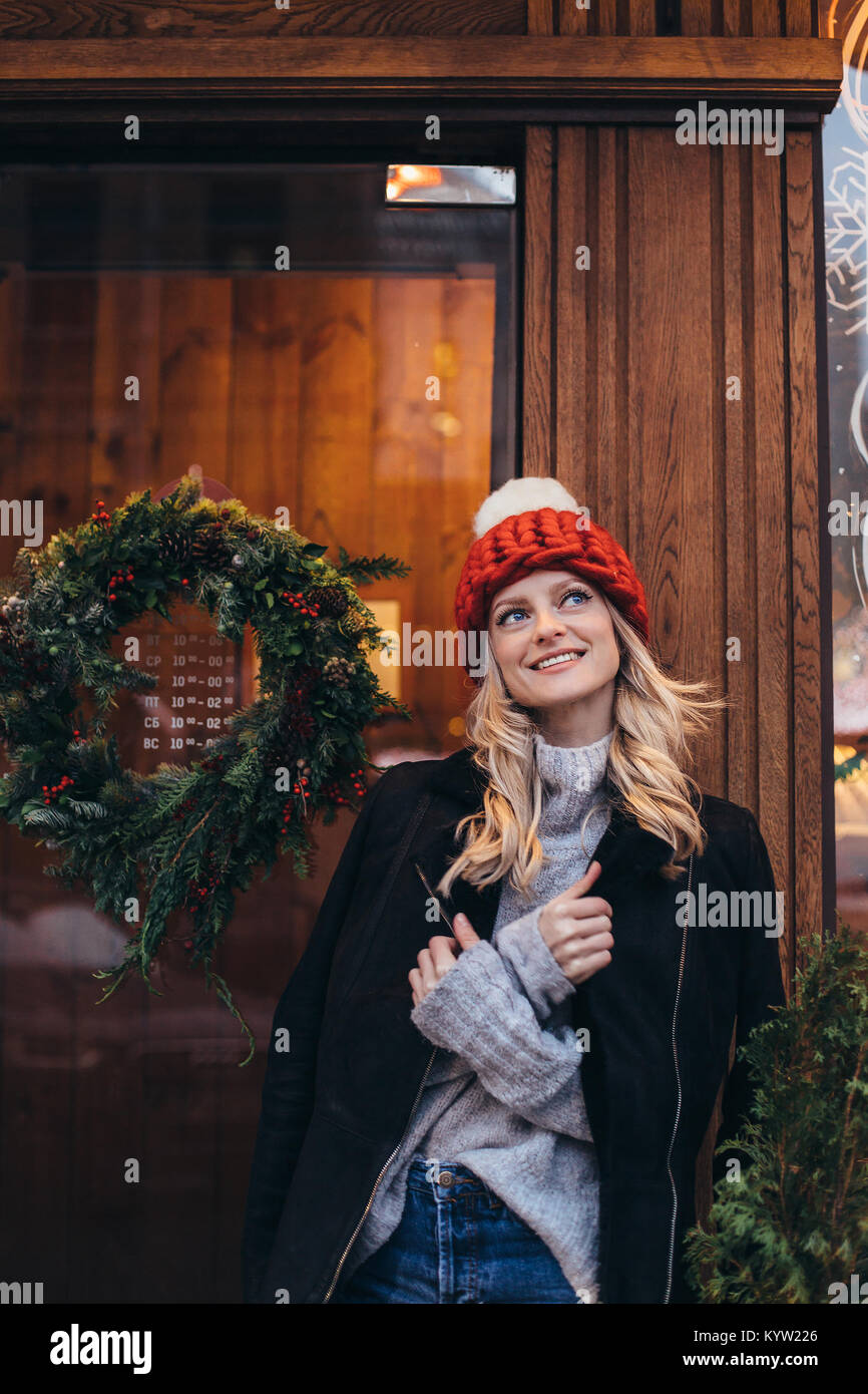 Young blonde woman in red hat tricoté avec pompon blanc, gris pull, veste noire et un jean bleu faire posant avec Christmac cafe fenêtre sur le backg Banque D'Images