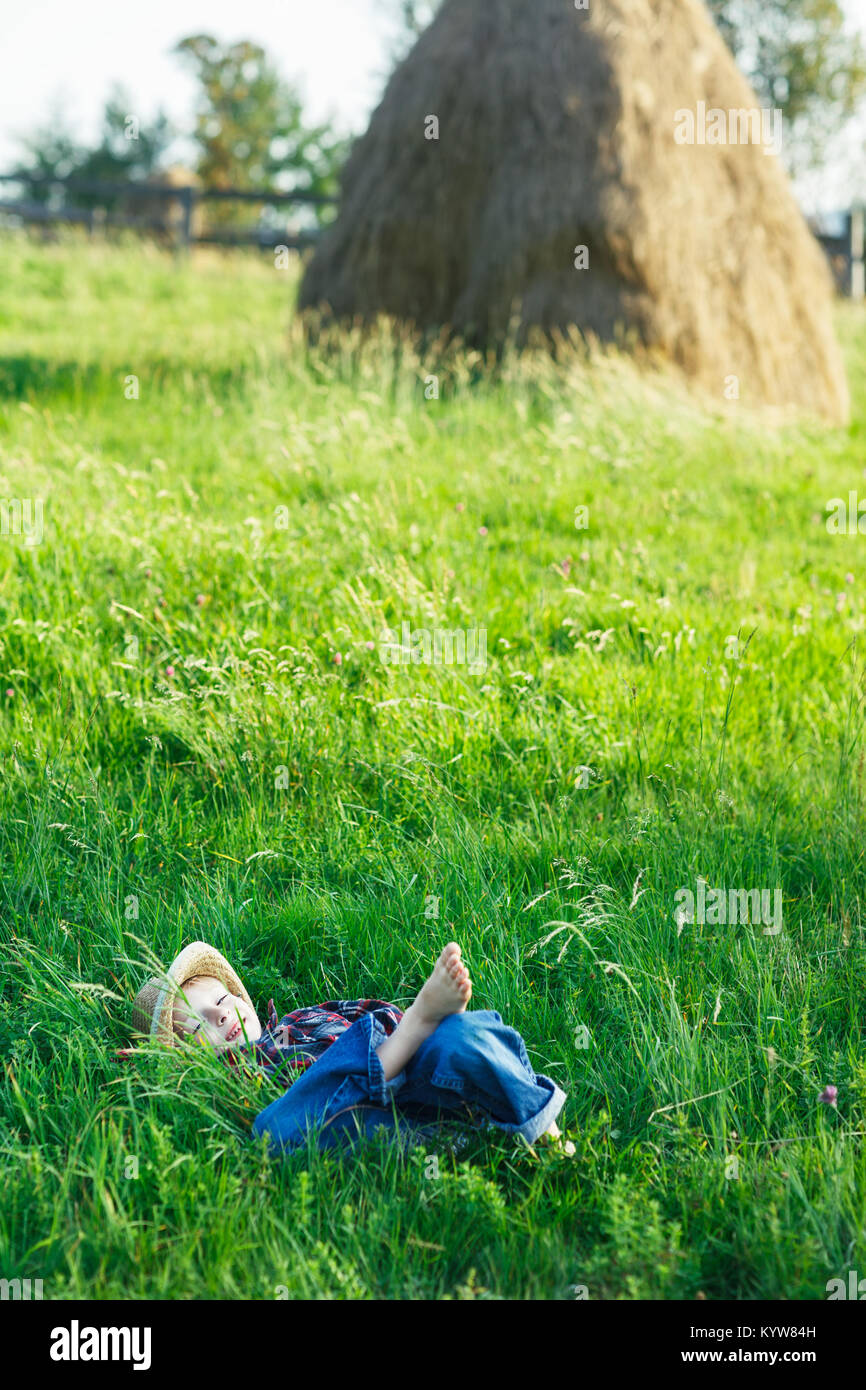 Joli petit garçon couché sur l'herbe sur le dos, les mains derrière la tête. Peu rêveur se trouve dans la région de prairie avec chapeau, les yeux fermés. Enfant endormi et souriant en natu Banque D'Images