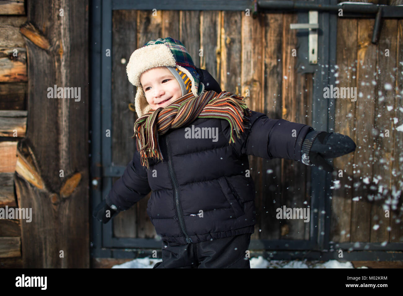 Smiling enfant debout contre le fond de bois avec la neige jetée en l'air. Garçon profitant de la neige. Banque D'Images