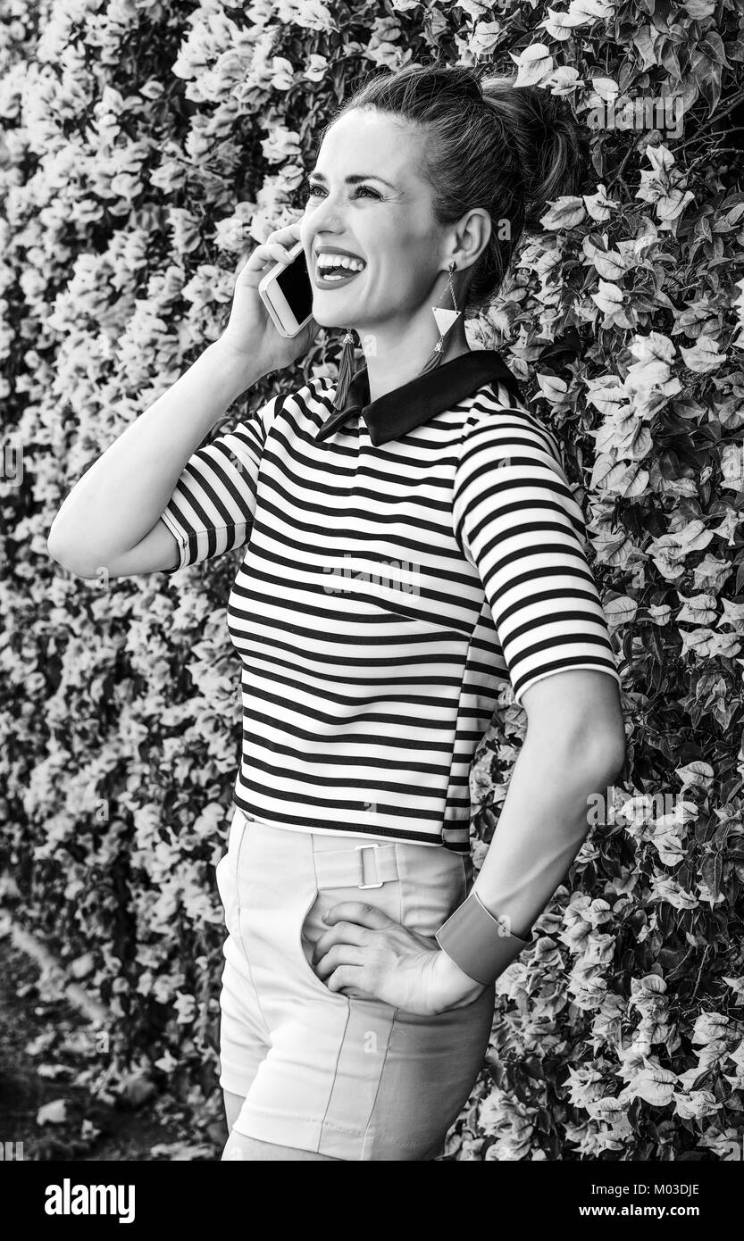 Fraîcheur colorée. smiling woman in yellow mode short et chemise à rayures à l'avant du lit coloré fleurs magenta parlant sur un téléphone cellulaire Banque D'Images