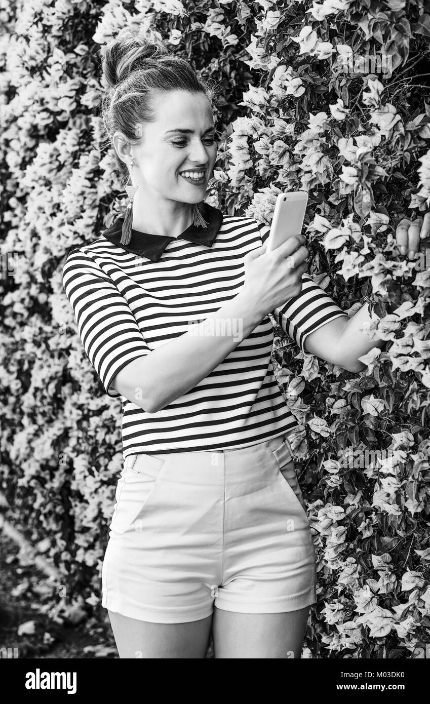 Fraîcheur colorée. smiling woman in élégant short jaune et stripy shirt against colorful fleurs magenta bed with cellphone taking photo Banque D'Images