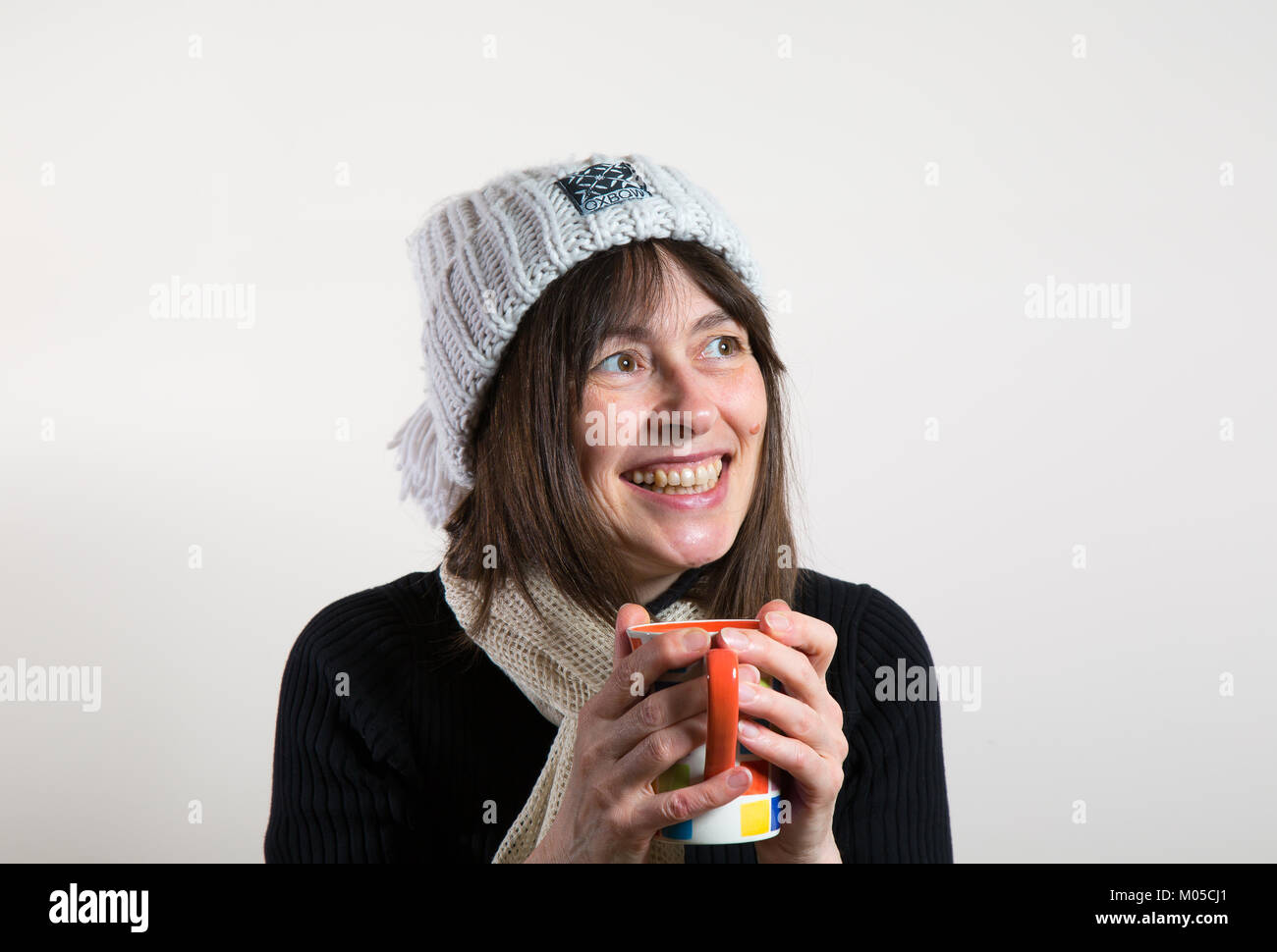 À l'intérieur portrait : happy female, vue avant, dans beanie bobble hat, tenant une tasse, un réchauffement de la main. Elle lève les yeux avec de grands sourire, sourire de fromage excité. Banque D'Images