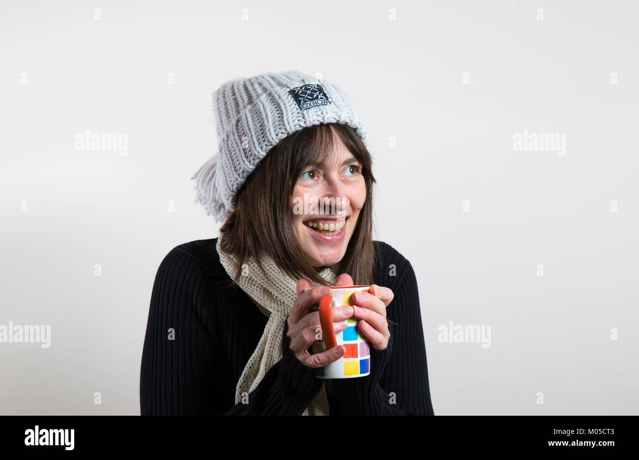 À l'intérieur portrait of happy girl in beanie bobble hat, tenant une tasse, un réchauffement de la main. Lève les yeux avec de grands sourire espiègle. Banque D'Images
