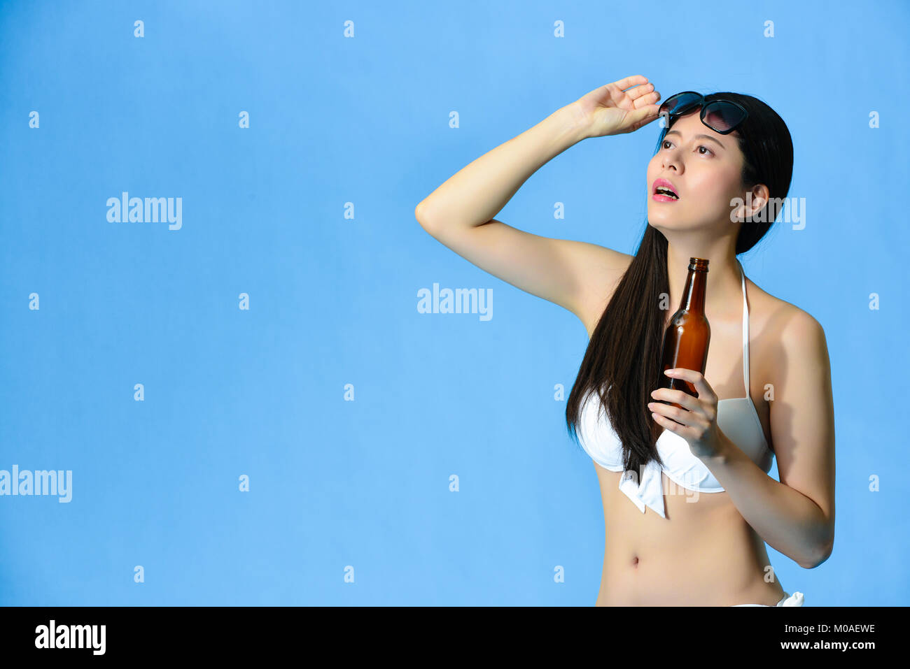 Malheureux jeune femme portant des vêtements de bikini de boire de la bière pendant la saison estivale et à la distance à avoir chaud isolé sur fond bleu. Banque D'Images
