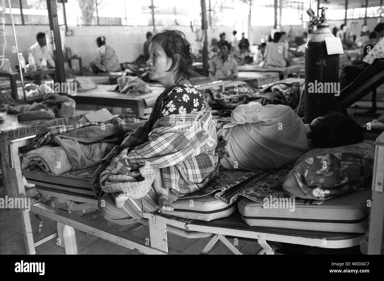 Une femme berceaux son bébé assis sur le lit de son mari gravement malade dans l'hospitalisation à l'unité clinique Mae Tao sur la frontière thaïlando-birmane. Environ 300 personnes par jour reçoivent des soins gratuits à la clinique, la plupart étant des travailleurs migrants et réfugiés qui ont franchi la frontière de la Birmanie (Myanmar) pour échapper à la pauvreté désespérée et la persécution du régime militaire. Les soins de santé sont tous mais inexistante en Birmanie, où moins de 2  % du PIB est consacré à la santé et l'éducation combinées. Banque D'Images