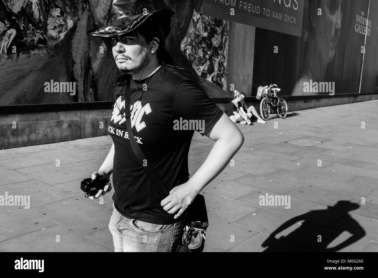 Photographie de rue en noir et blanc à Londres : Jeune homme regarde l'appareil photo sous la visière d'un chapeau en cuir en portant un T-shirt AC DC. Banque D'Images