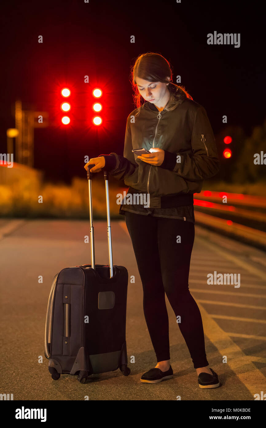 A young woman looking at smartphone lors de l'attente sur la plate-forme avec valise à la gare ; Georgetown, Ontario, Canada Banque D'Images