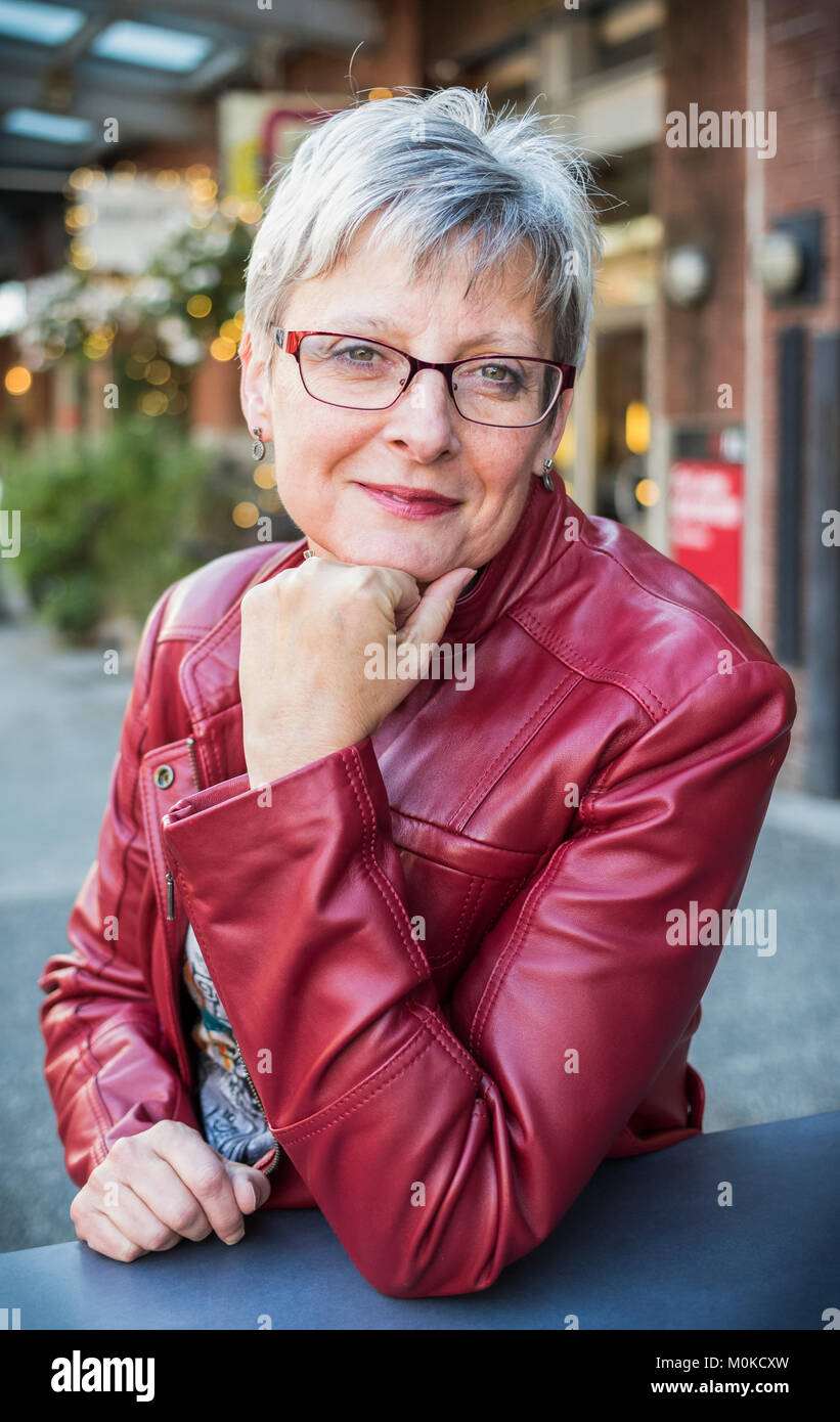 Portrait d'une femme mature dans une veste en cuir rouge assis à une table avec le menton reposant dans sa main ; Vancouver, Colombie-Britannique, Canada Banque D'Images