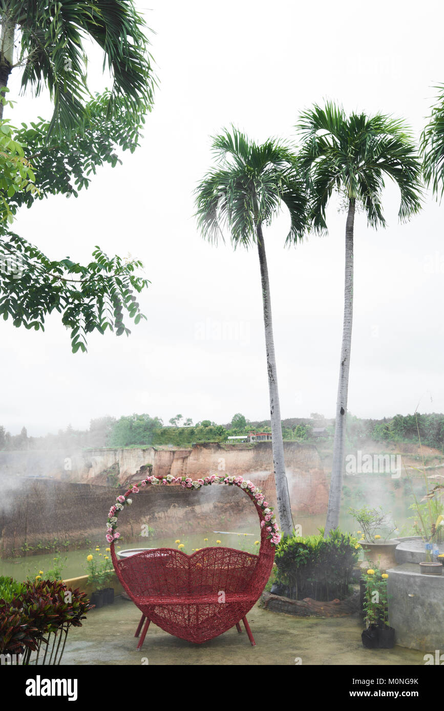 Banc avec un coeur rouge en forme décorée de fleurs contre un paysage tropical avec des palmiers en Thaïlande. Banque D'Images Banc avec un coeur rouge en forme décorée de fleurs contre un paysage tropical avec des palmiers en Thaïlande. Banque D'Images